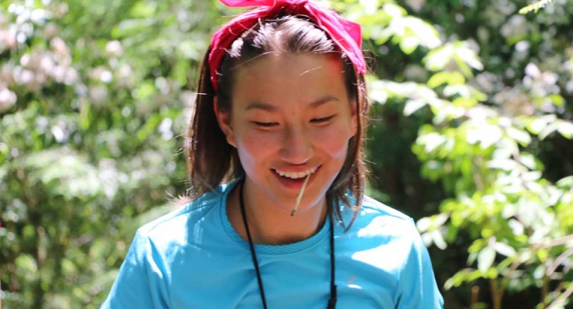 A young person smiles with a toothpick in their mouth. There are green leaves blurred behind them. 